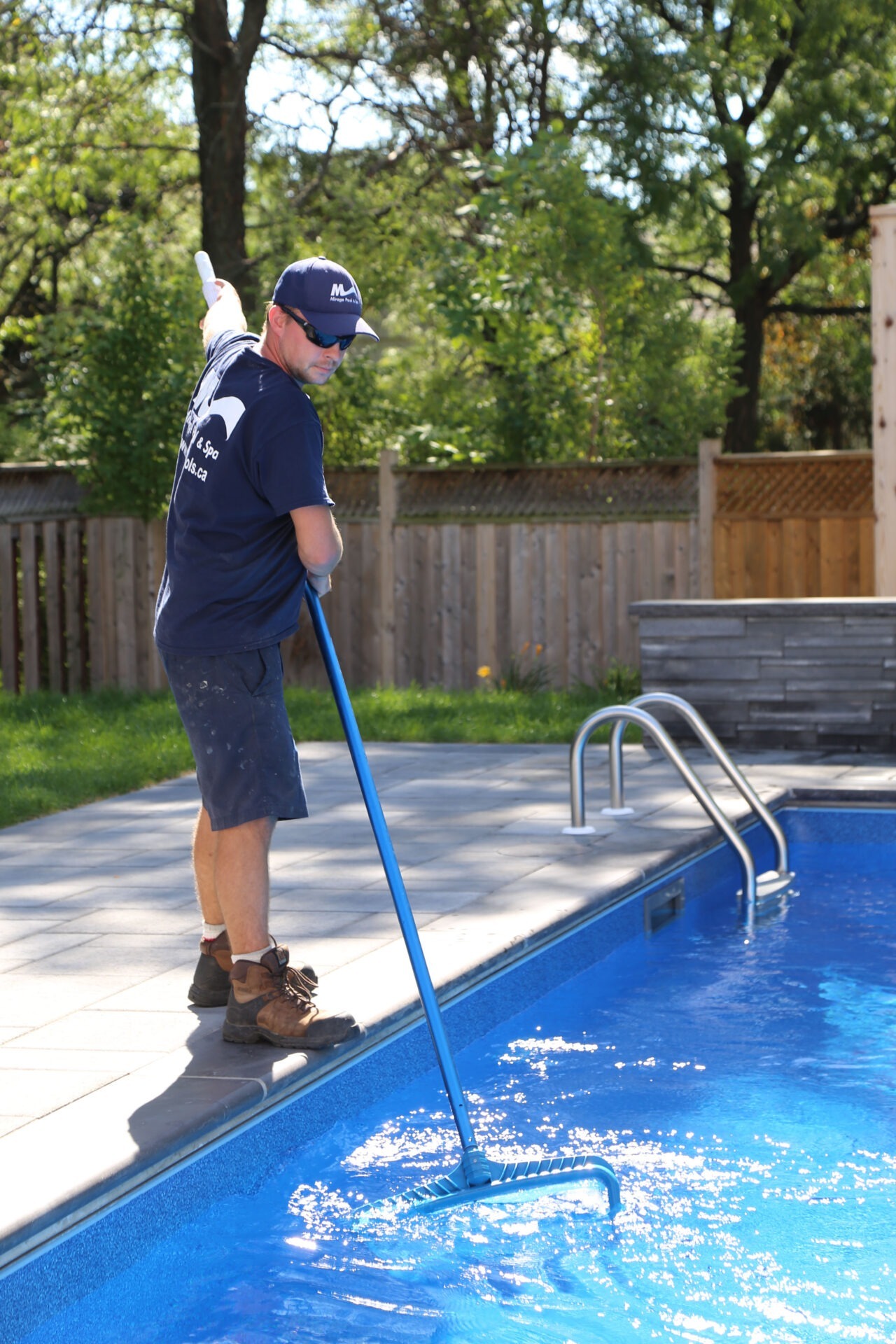 A person cleans a swimming pool with a skimmer in a backyard setting, surrounded by trees and a wooden fence.