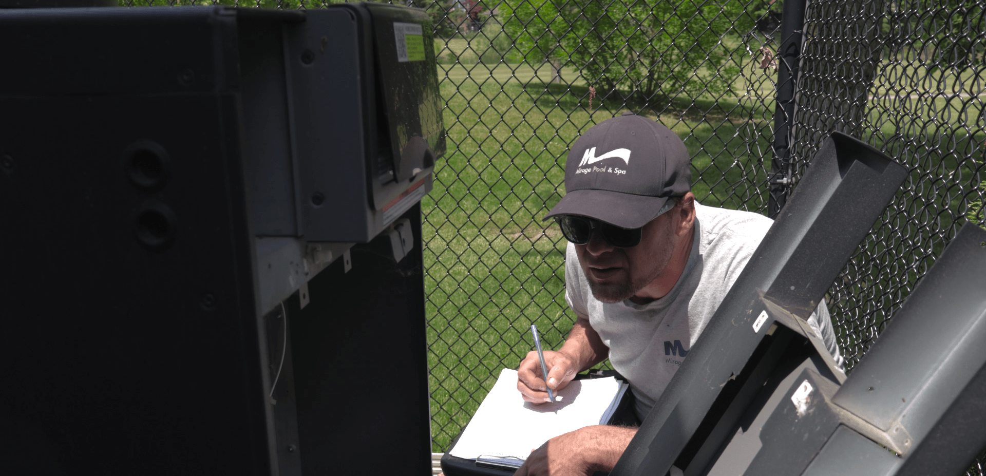 A person kneels by a large electrical box, taking notes on a clipboard, with a fenced grassy area in the background.