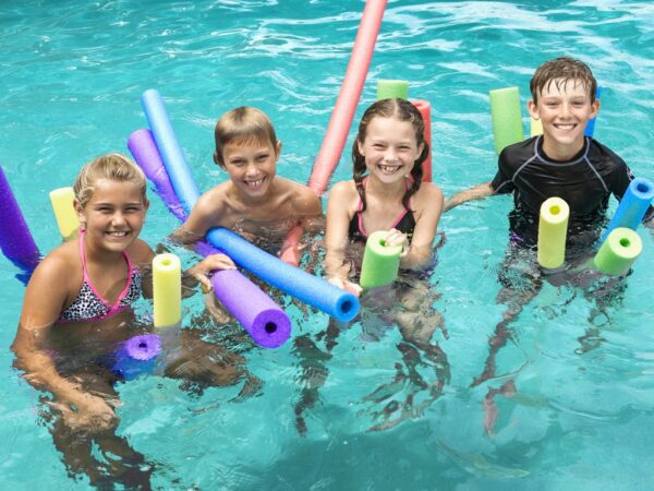 Four smiling children play in a pool using colorful pool noodles. The water is clear and the atmosphere is joyful and energetic.