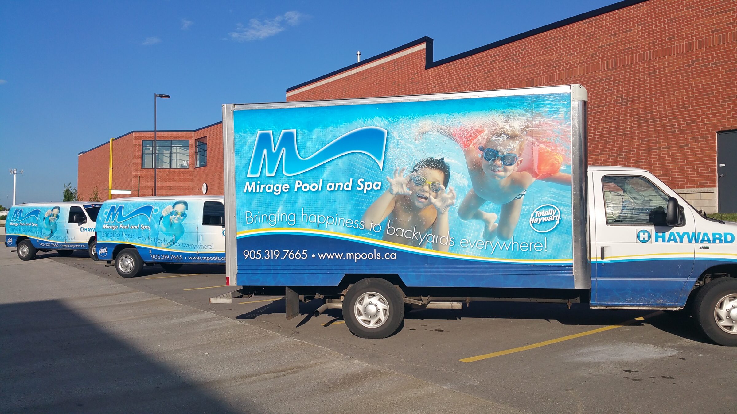 Three trucks with "Mirage Pool and Spa" branding are parked in a lot beside a large brick building. The sky is clear and sunny.