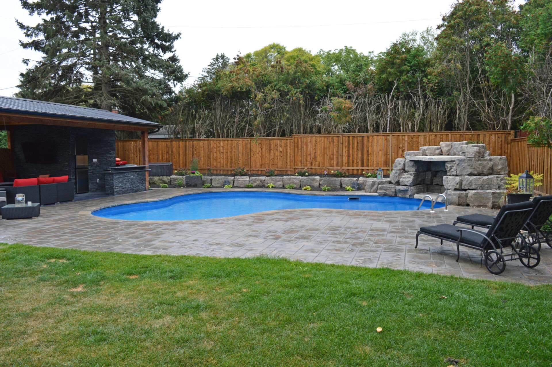 A backyard with a kidney-shaped pool, stone waterfall, lounge chairs, a gazebo with seating, a wooden fence, and surrounding greenery.