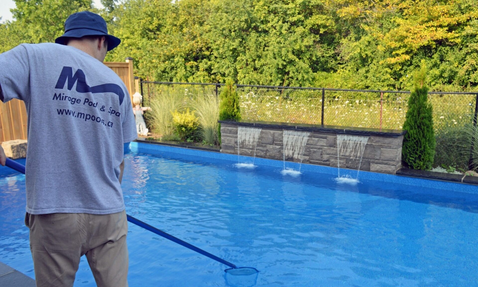 A person cleans a blue swimming pool with a net. Waterfalls cascade from a stone feature. Lush greenery surrounds the area.