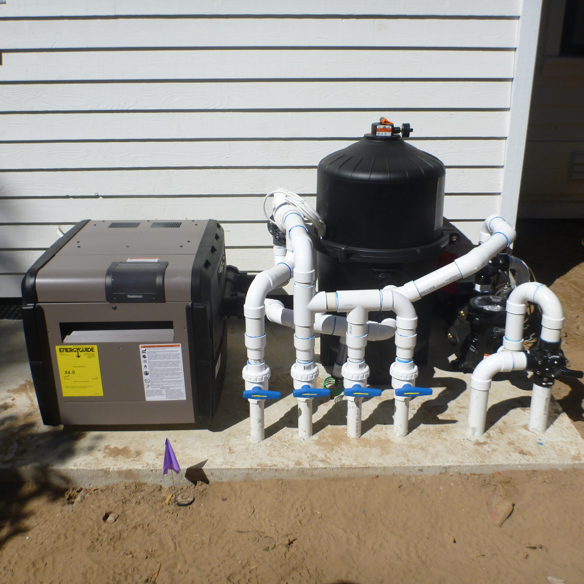 Pool equipment setup with a heater and filter system, featuring multiple PVC pipes and valves, placed outside a white building wall.