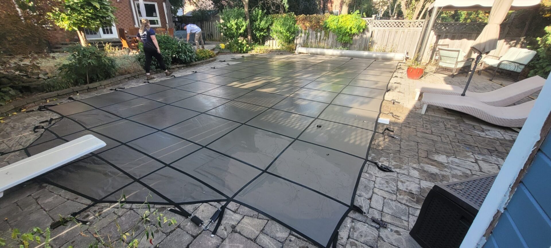 Two people secure a pool cover in a backyard with stone patio, surrounded by greenery, furniture, and a brick building.