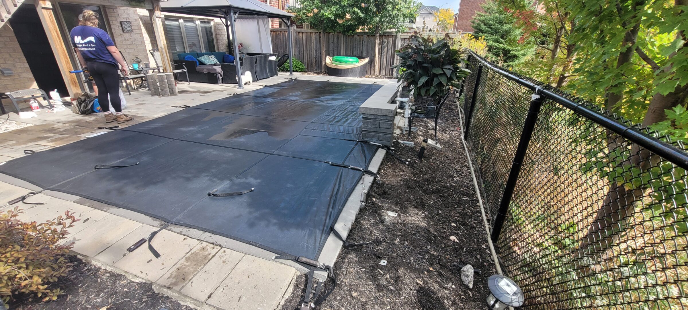 A person stands by a covered pool in a backyard with patio furniture, greenery, and a chain-link fence.