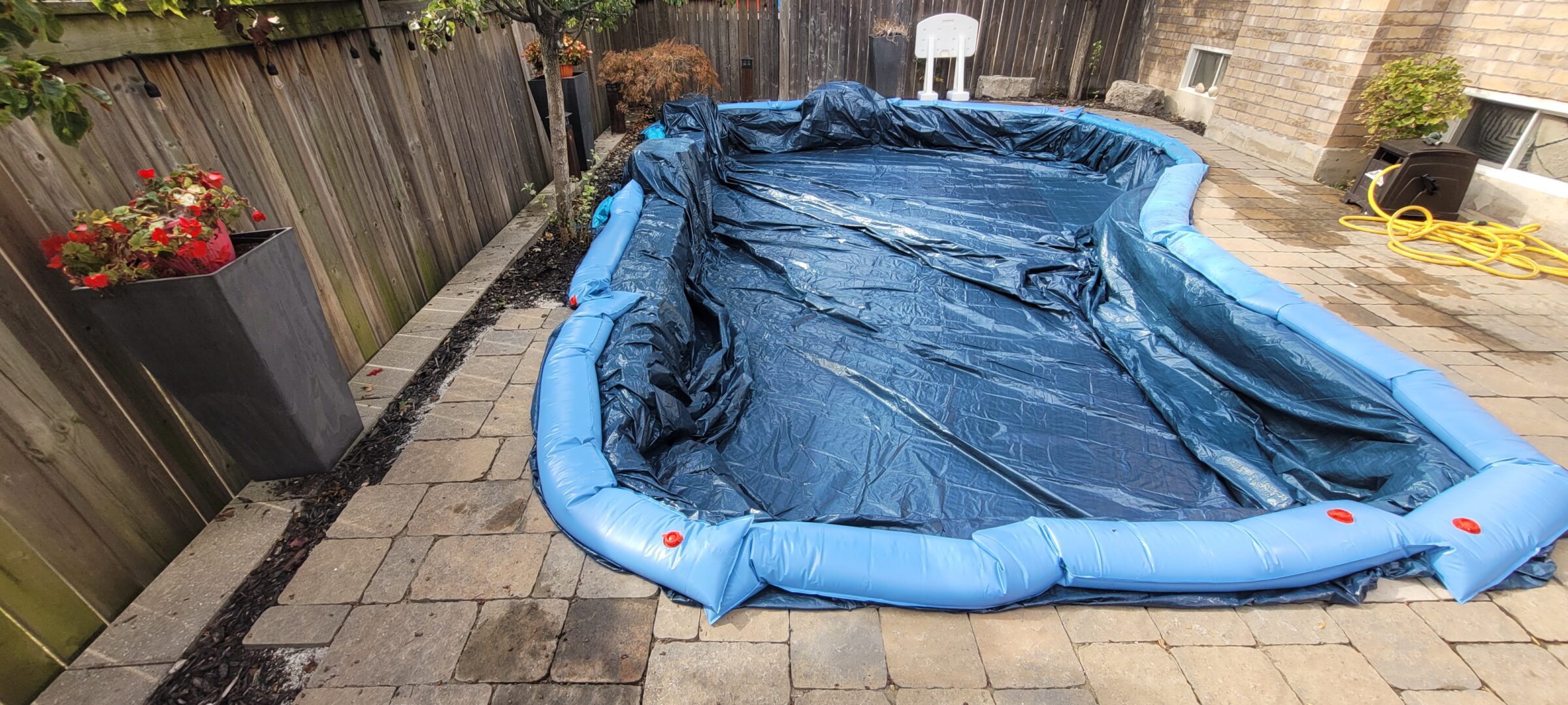 An empty above-ground pool covered with a blue tarp in a fenced backyard, surrounded by potted plants on a stone patio.