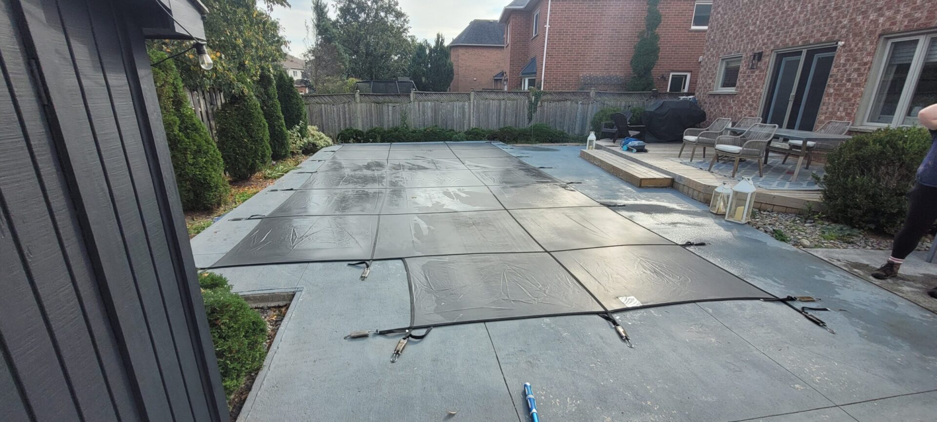 A backyard with a covered pool, patio area, dining set, and person. Brick houses and trees surround the space, creating a cozy atmosphere.