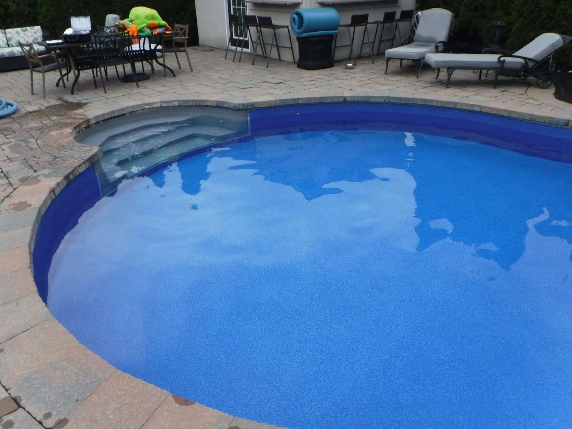 A backyard scene with a blue swimming pool, surrounded by patio furniture, lounge chairs, and a barbecue grill on a stone deck.