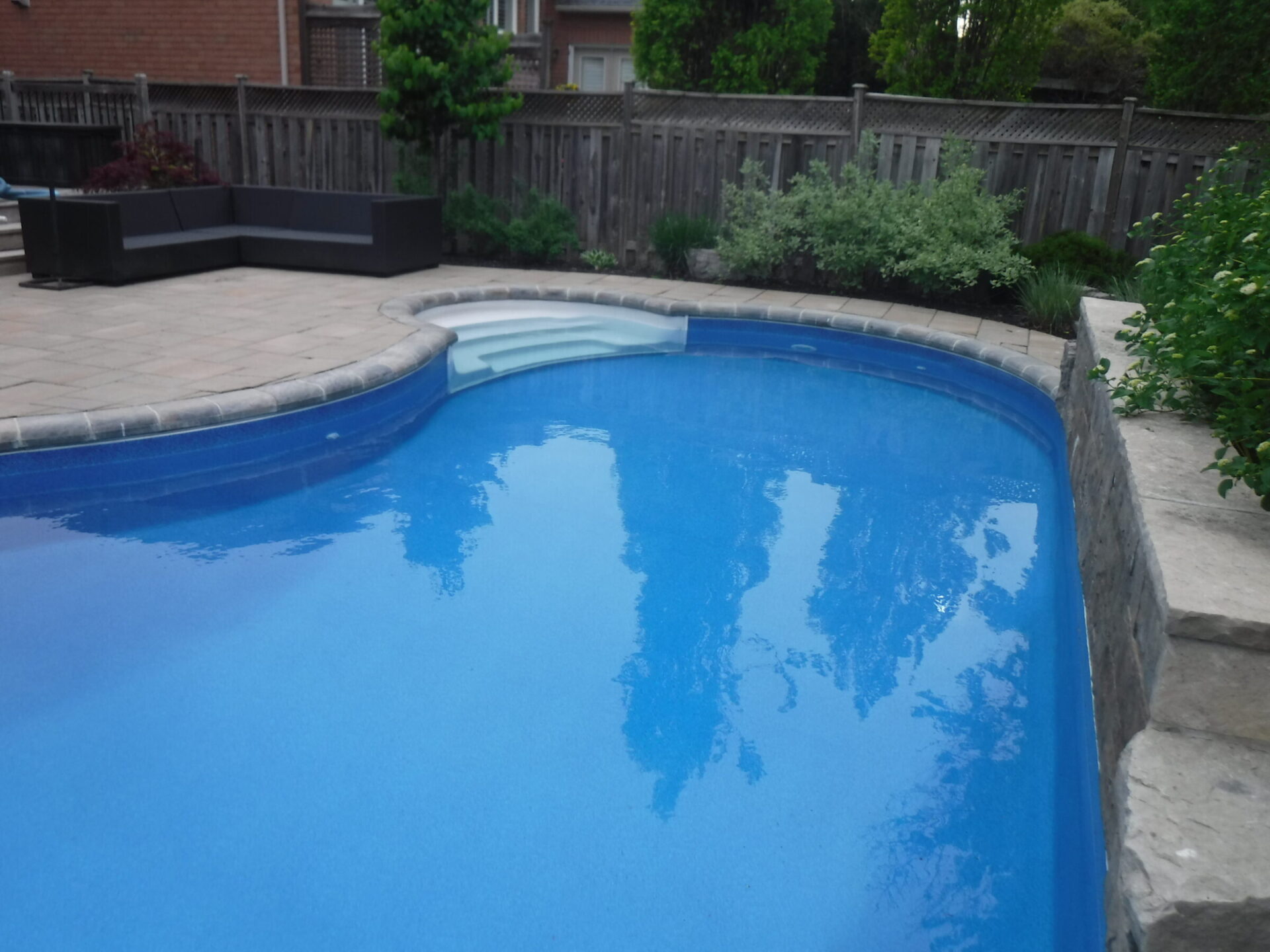 A tranquil backyard scene featuring a clear blue swimming pool, patio seating area, and lush greenery enclosed by a wooden fence.