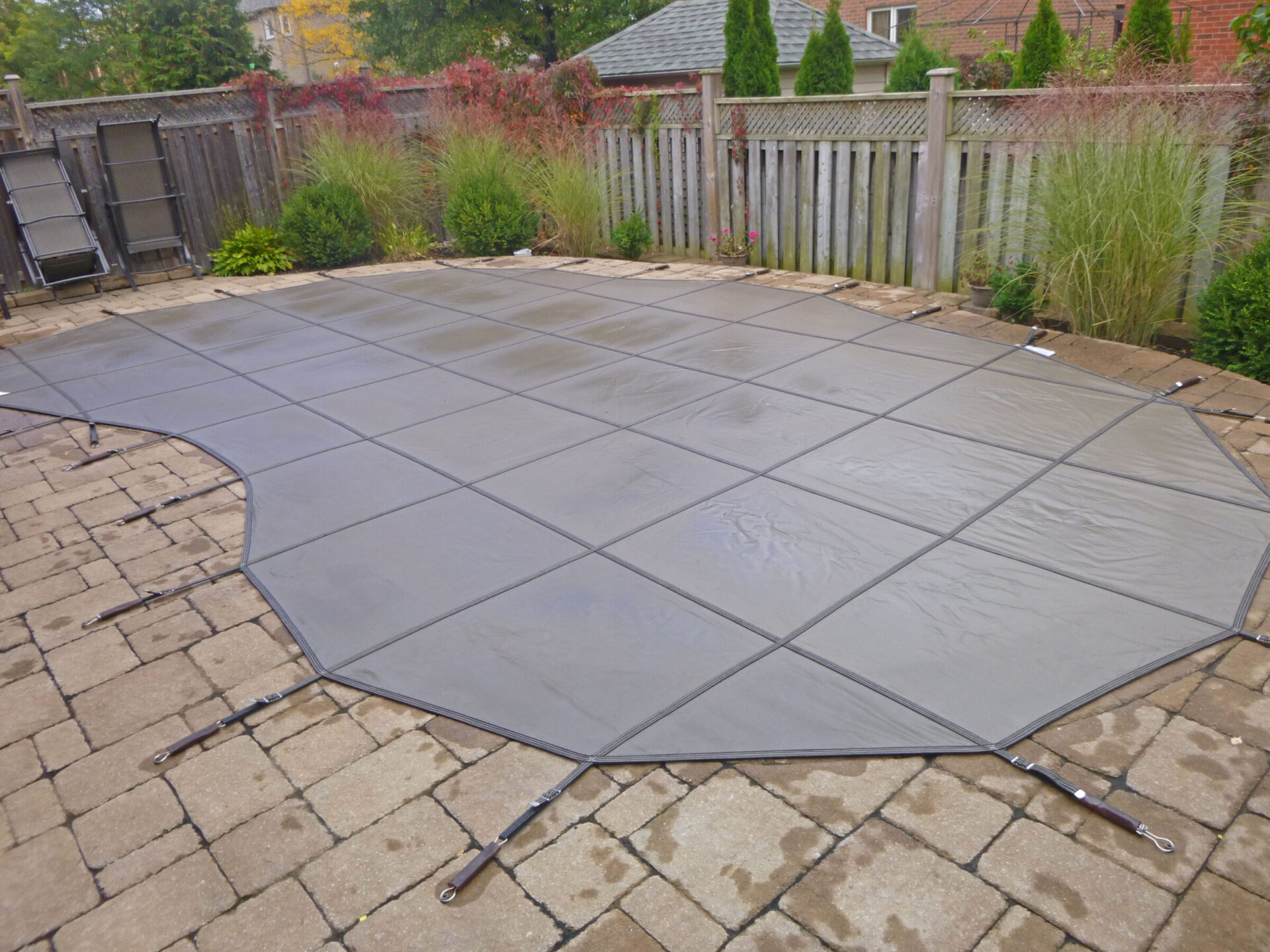 A backyard with a closed pool covered by a large tarp, surrounded by a wooden fence and tall plants on a stone patio.