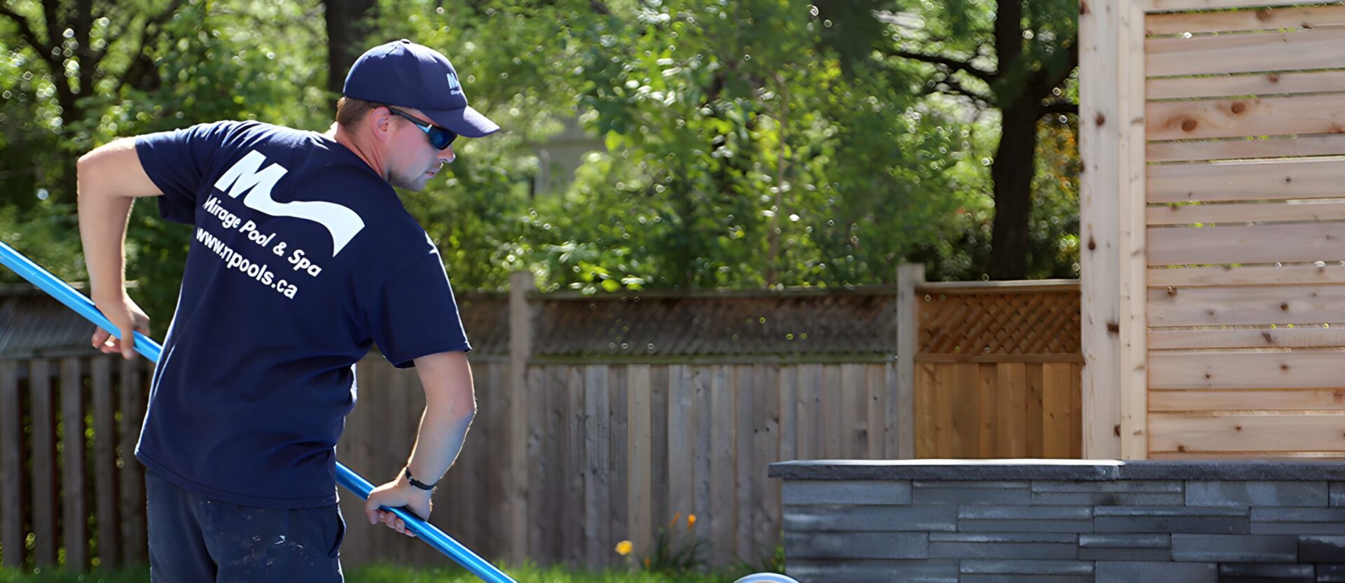 A person wearing a blue uniform and cap is cleaning a pool with a pole beside a wooden fence and trees.