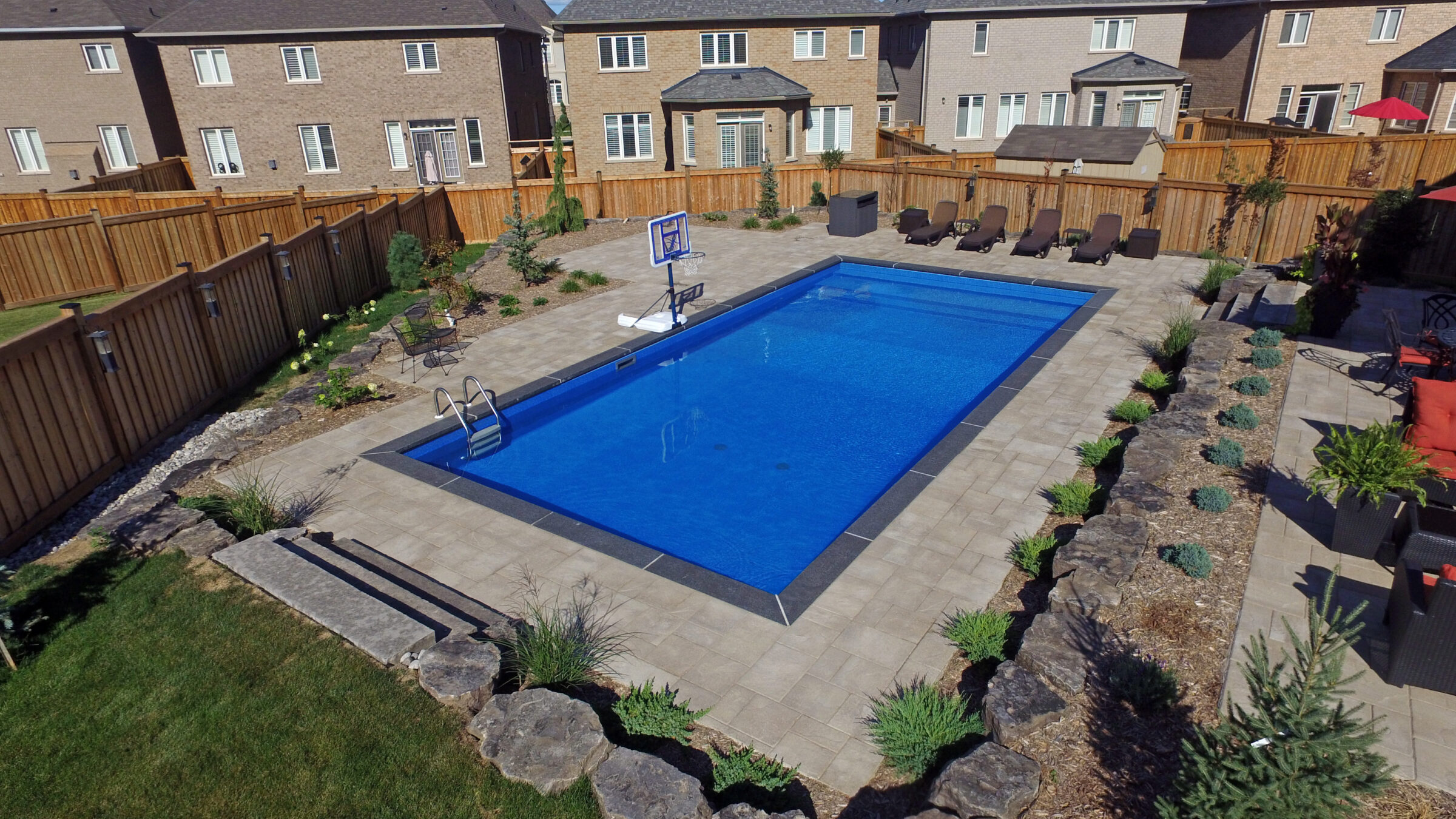 A private backyard features a rectangular swimming pool, surrounded by stone paving, a basketball hoop, lounge chairs, and a wooden fence.