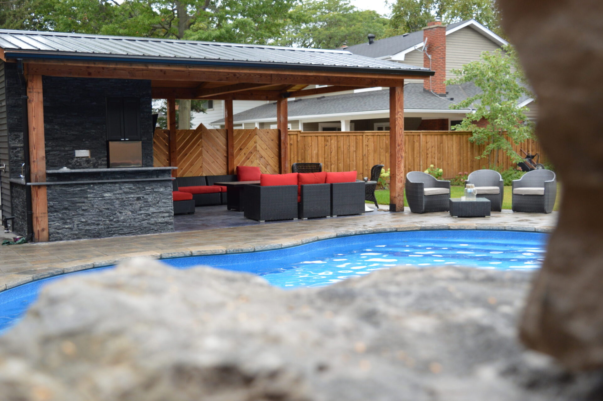 A backyard scene features a swimming pool, a covered outdoor lounge area with red and black furniture, and a wooden fence in the background.