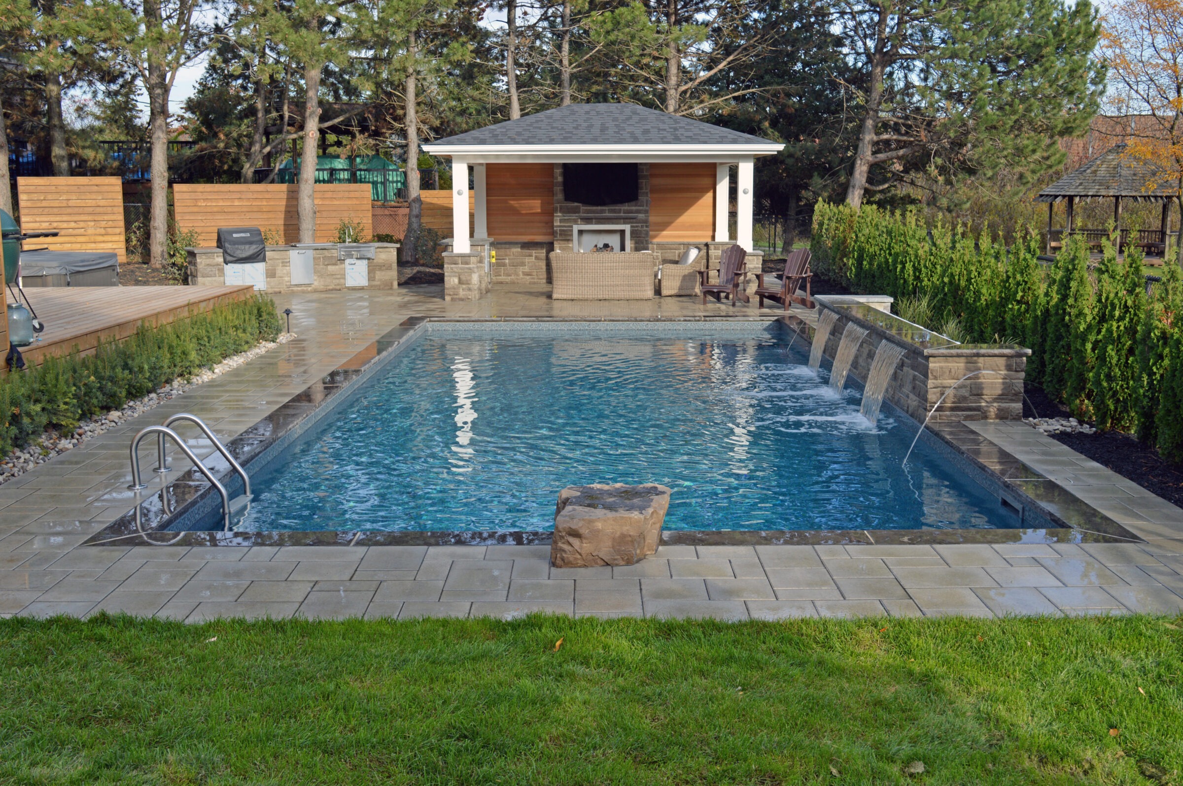 A backyard features a rectangular pool with fountain jets, surrounded by stone paving, greenery, and a covered seating area under a gazebo.