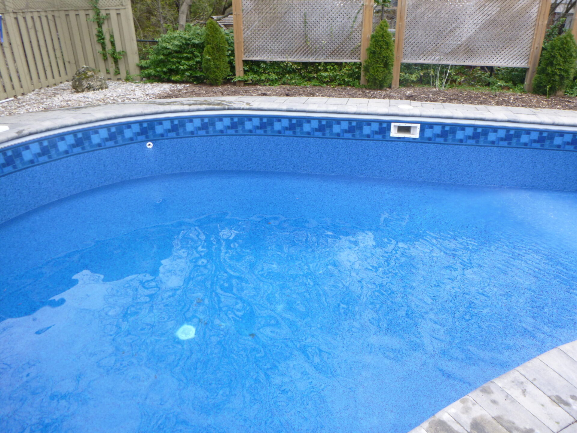 A backyard swimming pool with clear blue water is surrounded by stone tiles and wooden fencing, accented with small plants and shrubs.