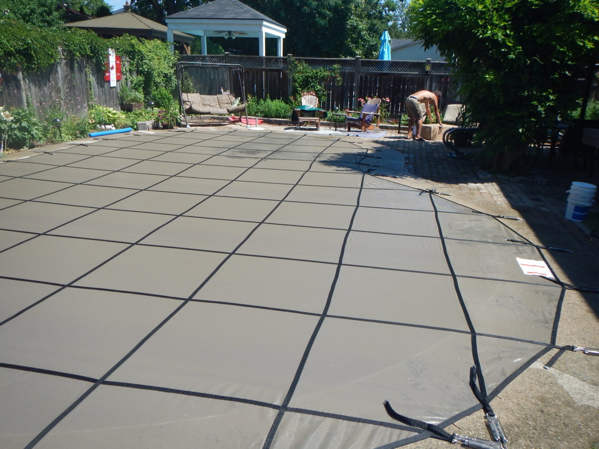 A fenced backyard with a covered pool and patio furniture. Two people are nearby, one standing by the pool. Trees and shrubs surround the area.