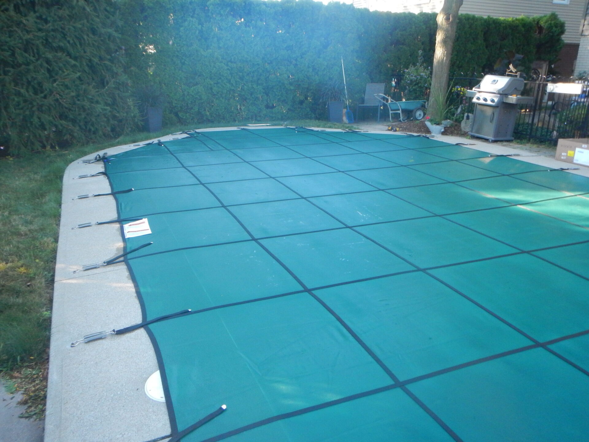 A backyard scene featuring a pool covered with a green tarp, surrounded by trees, patio furniture, and a barbecue grill.