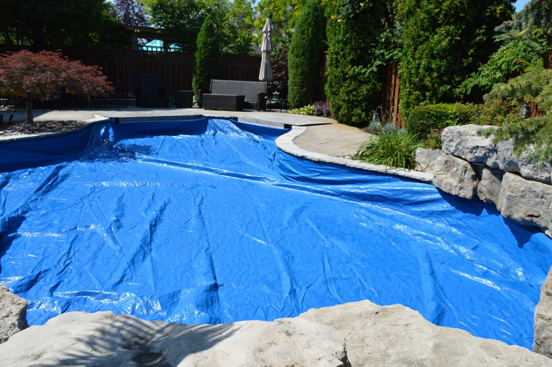 A backyard scene with a pool covered in a blue tarp, surrounded by rocks and greenery; patio furniture seen in the background.