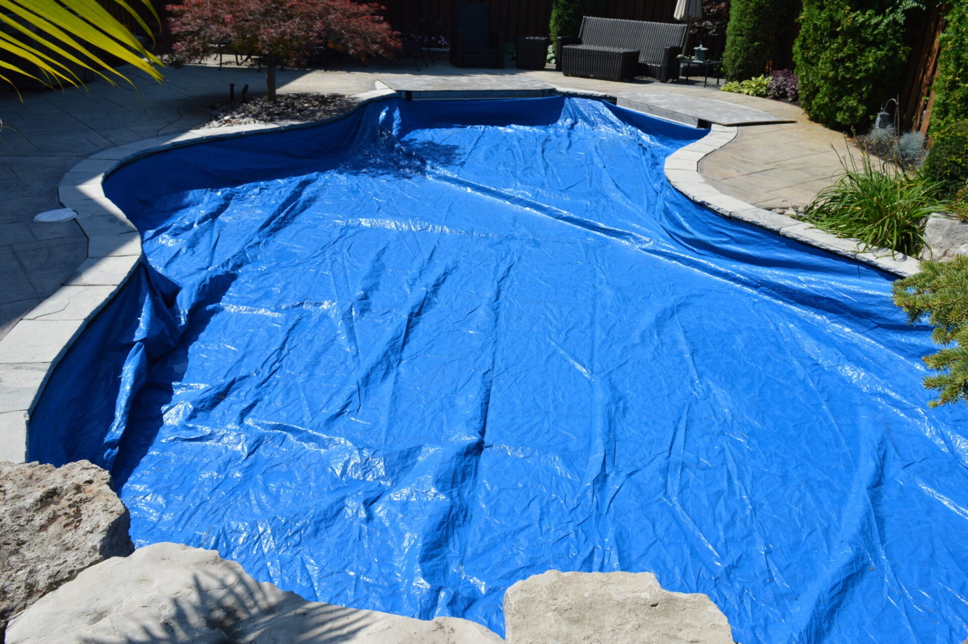 A backyard pool is covered with a large blue tarp. Surrounding landscaping includes shrubs, a rock border, and patio furniture in the background.