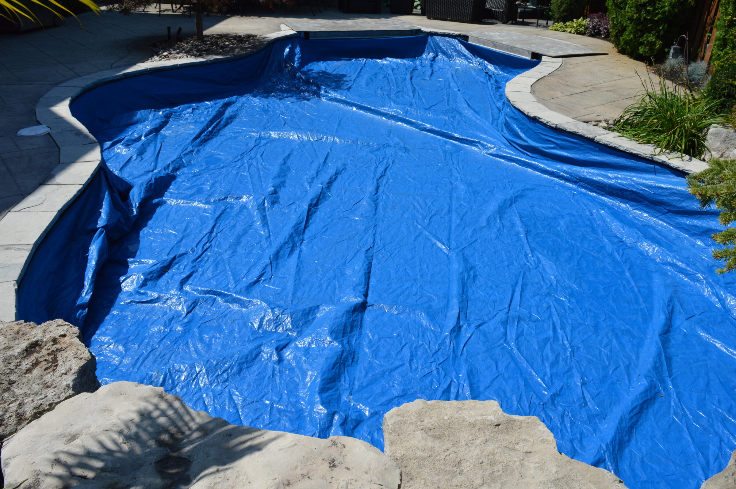 A swimming pool covered with a blue tarp surrounded by stone and plants. No people or recognizable landmarks are visible.