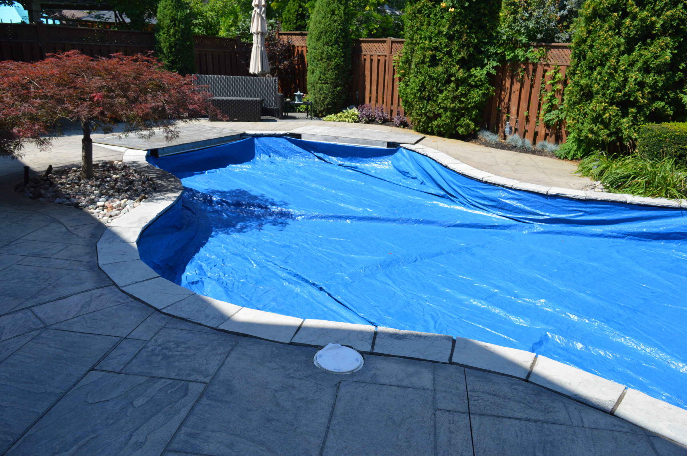 Backyard with a partially covered pool surrounded by concrete paving, a small tree, and privacy fencing. Greenery and patio furniture in the background.