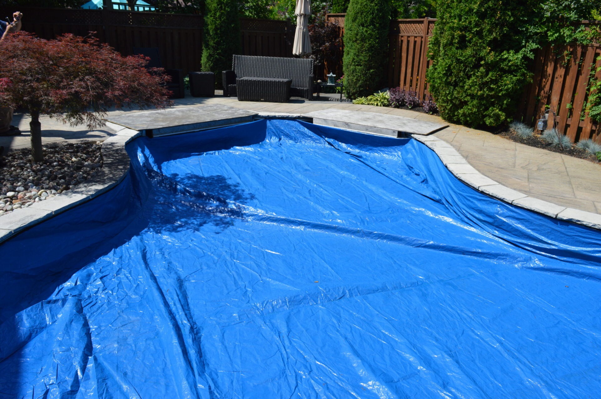 A backyard with a swimming pool covered by a blue tarp, surrounded by a wooden fence, patio furniture, and trees.