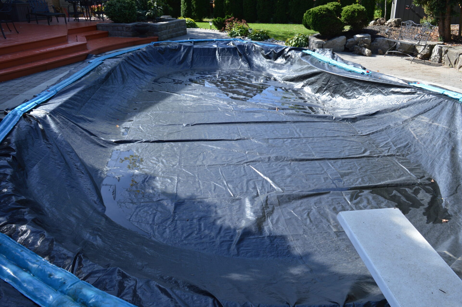 A backyard swimming pool is covered with a black tarp. Nearby are red brick steps, greenery, and a diving board.