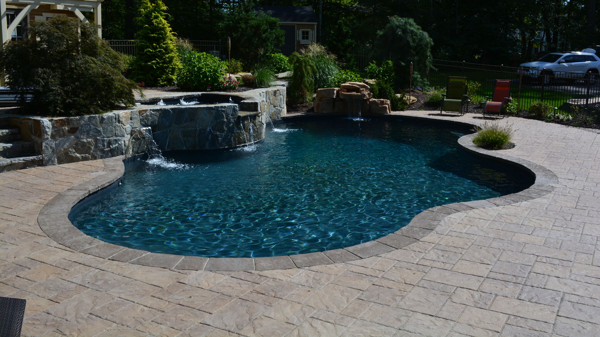 A serene backyard pool surrounded by stone, lush greenery, and lounge chairs under a sunny sky, with a car visible in the background.