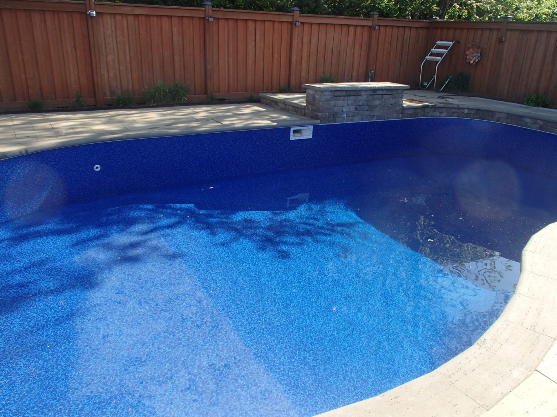 This image shows an empty backyard pool with a blue liner, surrounded by a wooden fence and a paved area with a small platform.