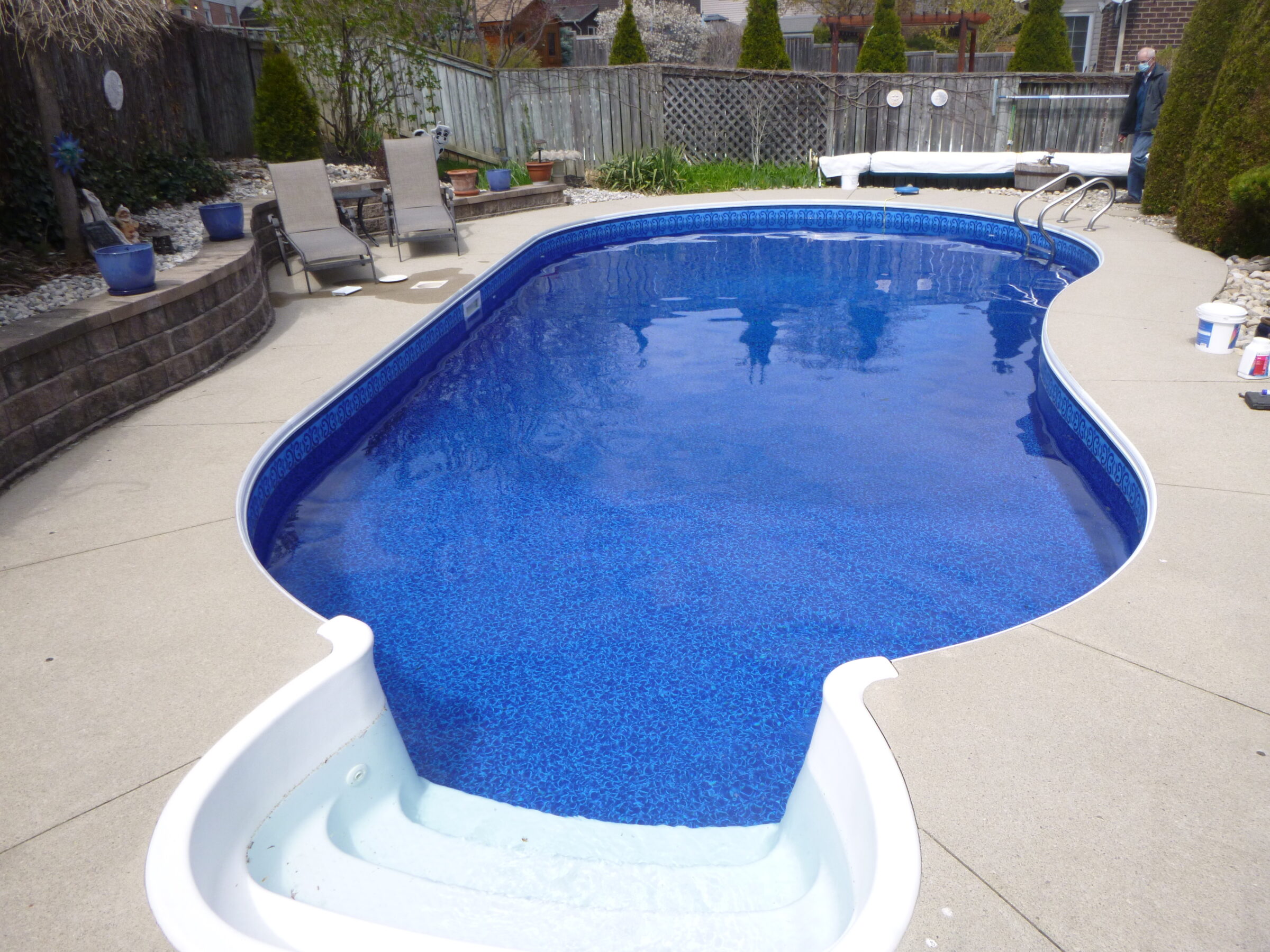 A backyard features an in-ground swimming pool with curved edges, surrounded by lounge chairs and a wooden fence. No people are visible.