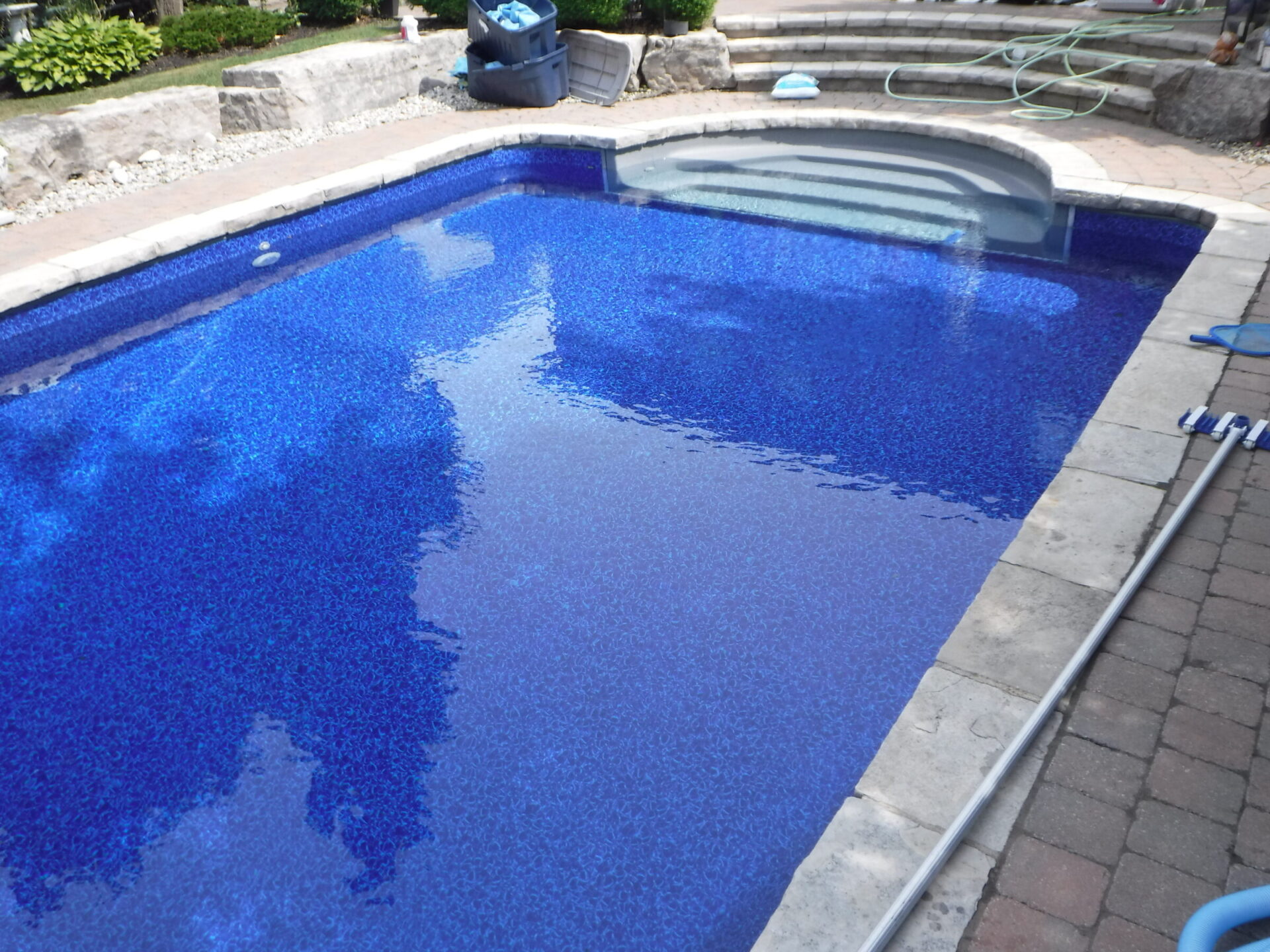 A clear blue swimming pool with steps, surrounded by stone paving. Gardening tools and a hose are visible nearby. No landmarks present.