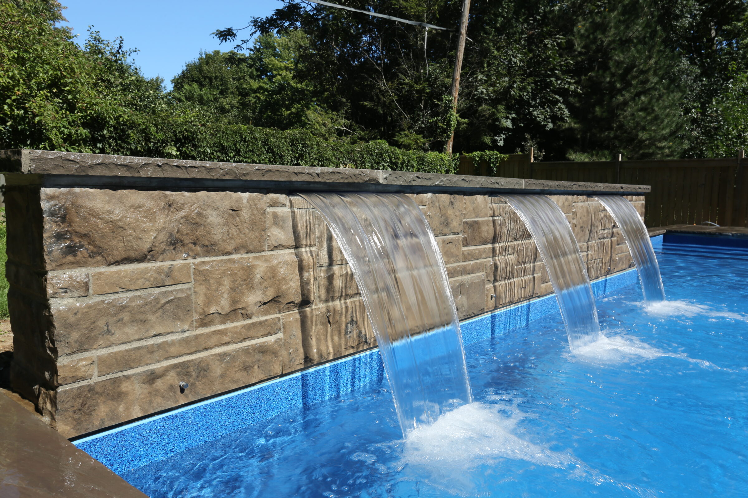 A stone wall with cascading waterfalls overlooks a clear blue pool, surrounded by lush green trees and a wooden fence.