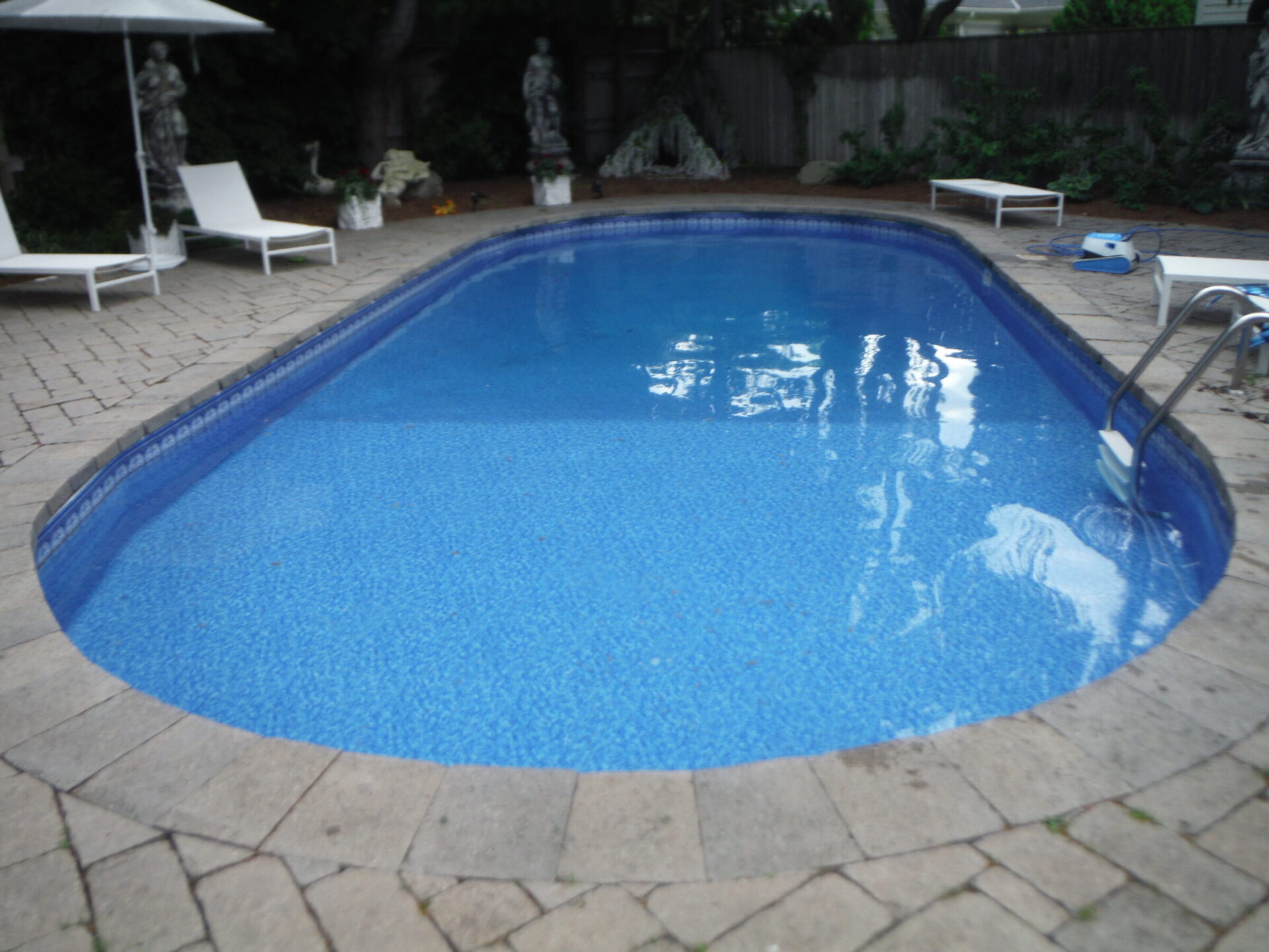 A serene backyard pool surrounded by lounge chairs, statues, and trees, with a stone patio. No people or notable landmarks visible.