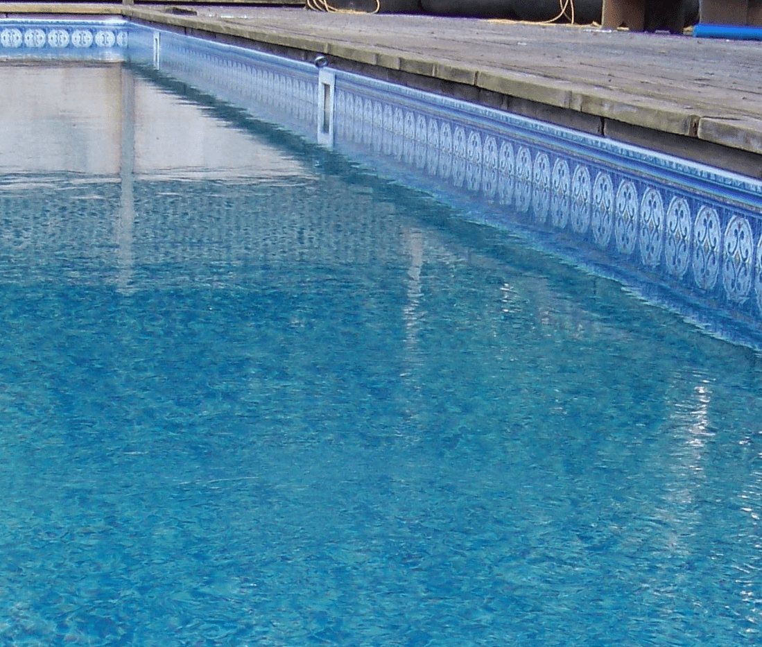 A swimming pool with clear blue water and patterned tiles, adjacent to a wooden deck. No people or landmarks visible.