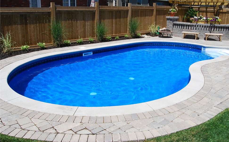 A backyard features an oval swimming pool surrounded by stone pavers, with wooden fencing, plants, and benches in the background.