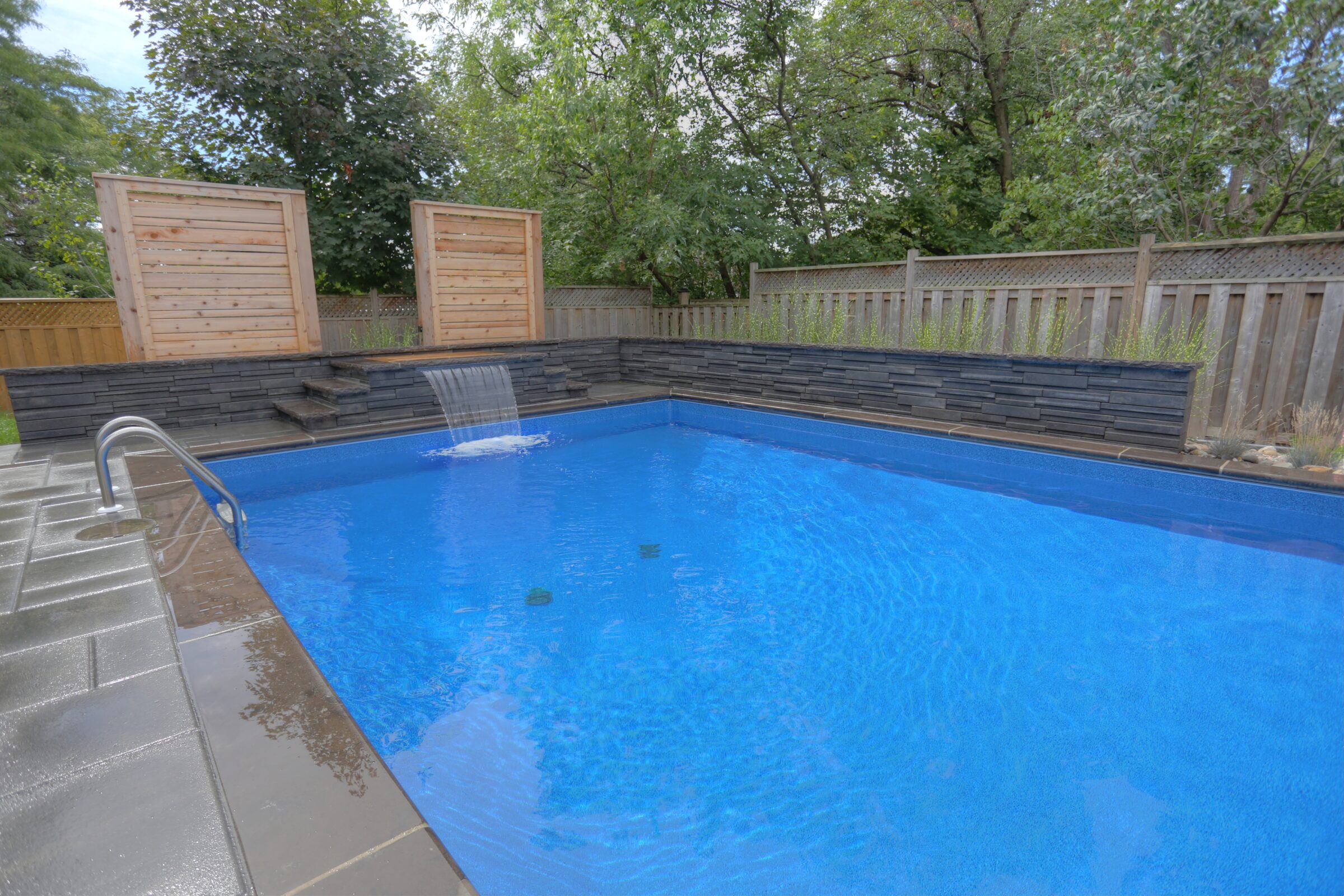 A serene backyard pool with a waterfall feature, surrounded by wooden fencing and lush greenery, under a clear blue sky.