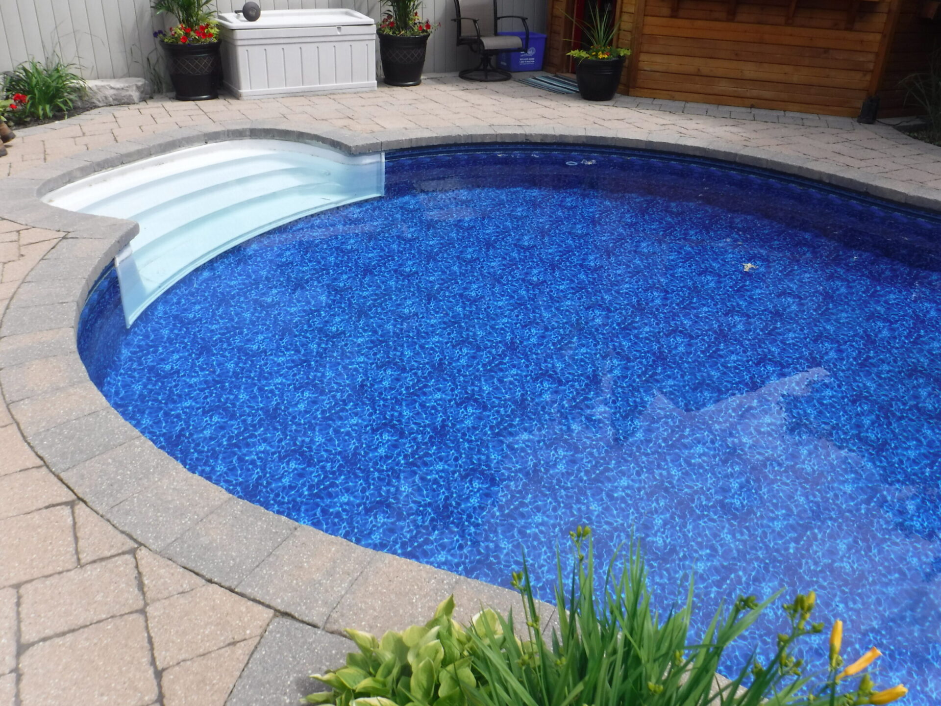 A small, blue swimming pool with white steps is surrounded by a stone patio. Potted plants and flowers decorate the area.