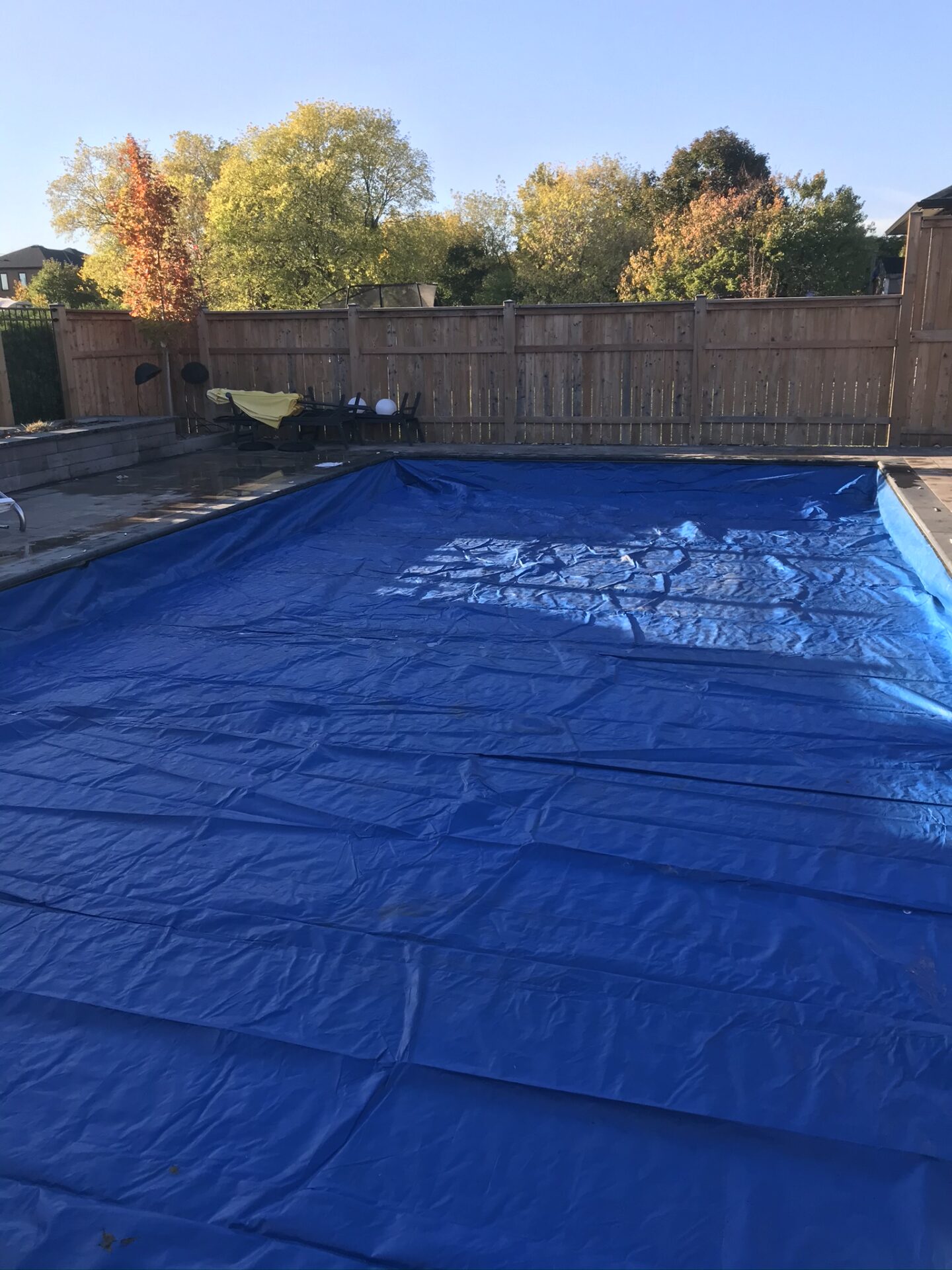 A backyard pool covered with a blue tarp, surrounded by a wooden fence, patio furniture, and trees with autumn foliage.