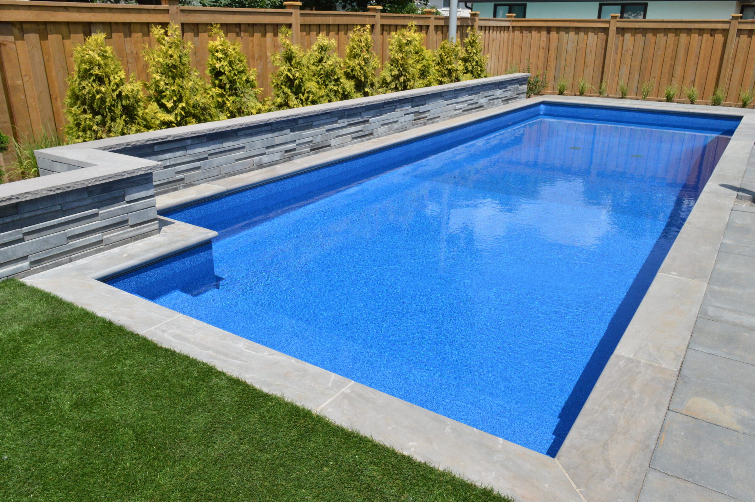 A rectangular swimming pool with clear blue water, surrounded by stone tiles and a wooden fence, with greenery along the edges.