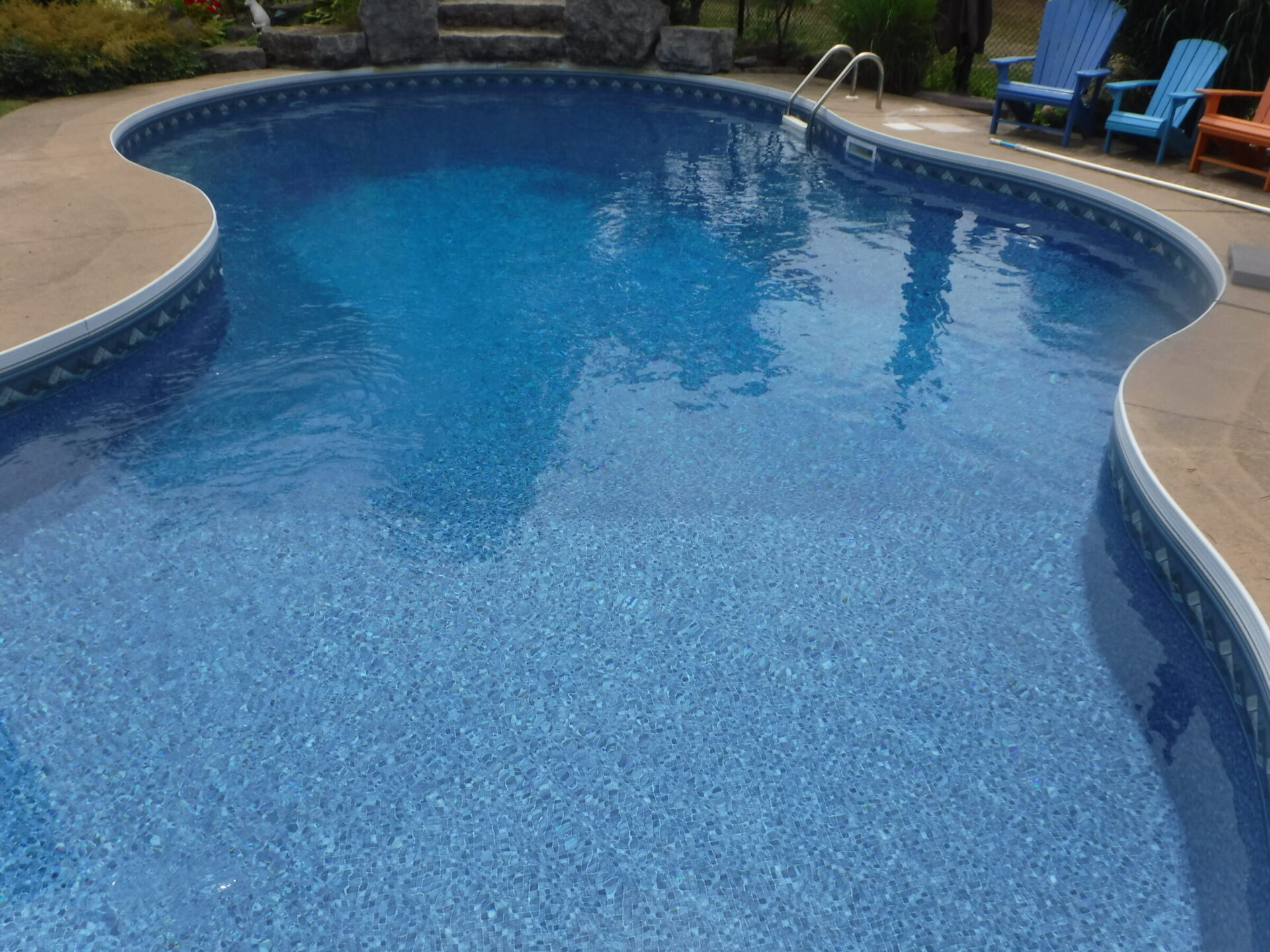 A serene pool with clear blue water, surrounded by patio chairs. Lush greenery frames the area, creating a tranquil outdoor setting.