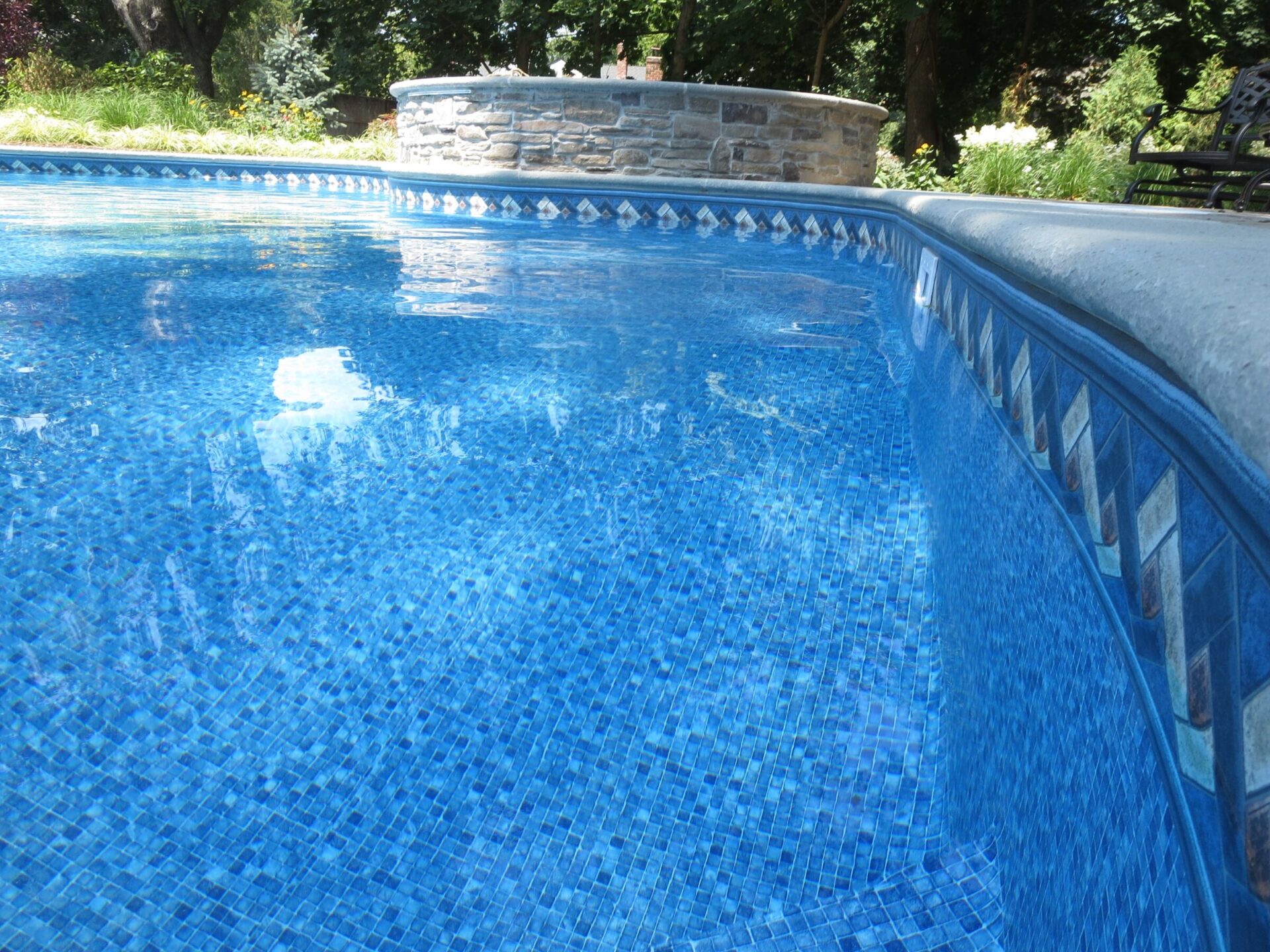 A swimming pool with blue mosaic tiles, surrounded by lush greenery. A stone structure is visible in the background under a clear sky.