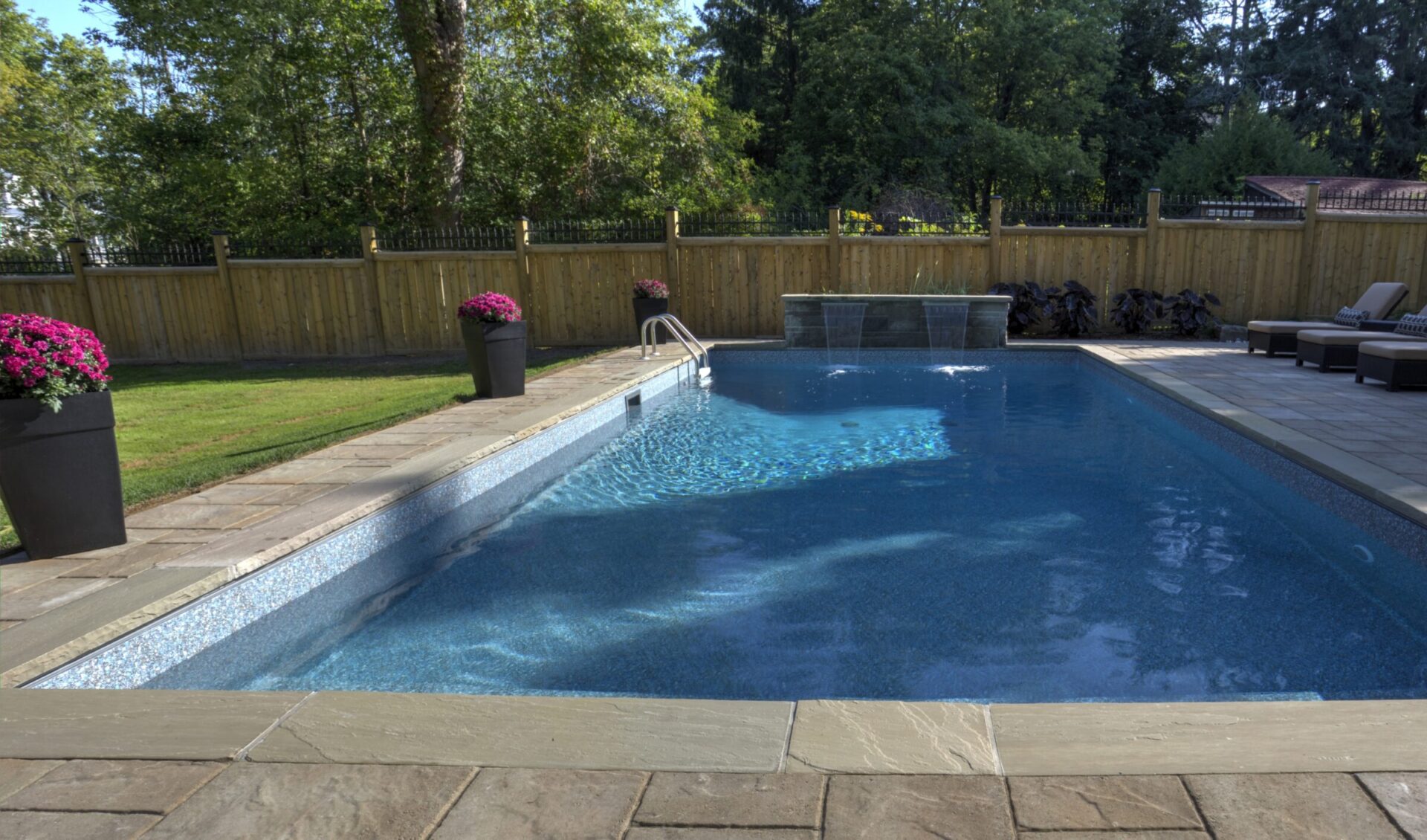 A pristine backyard pool with stone decking, surrounded by a wooden fence, greenery, and potted flowers. No people or landmarks visible.