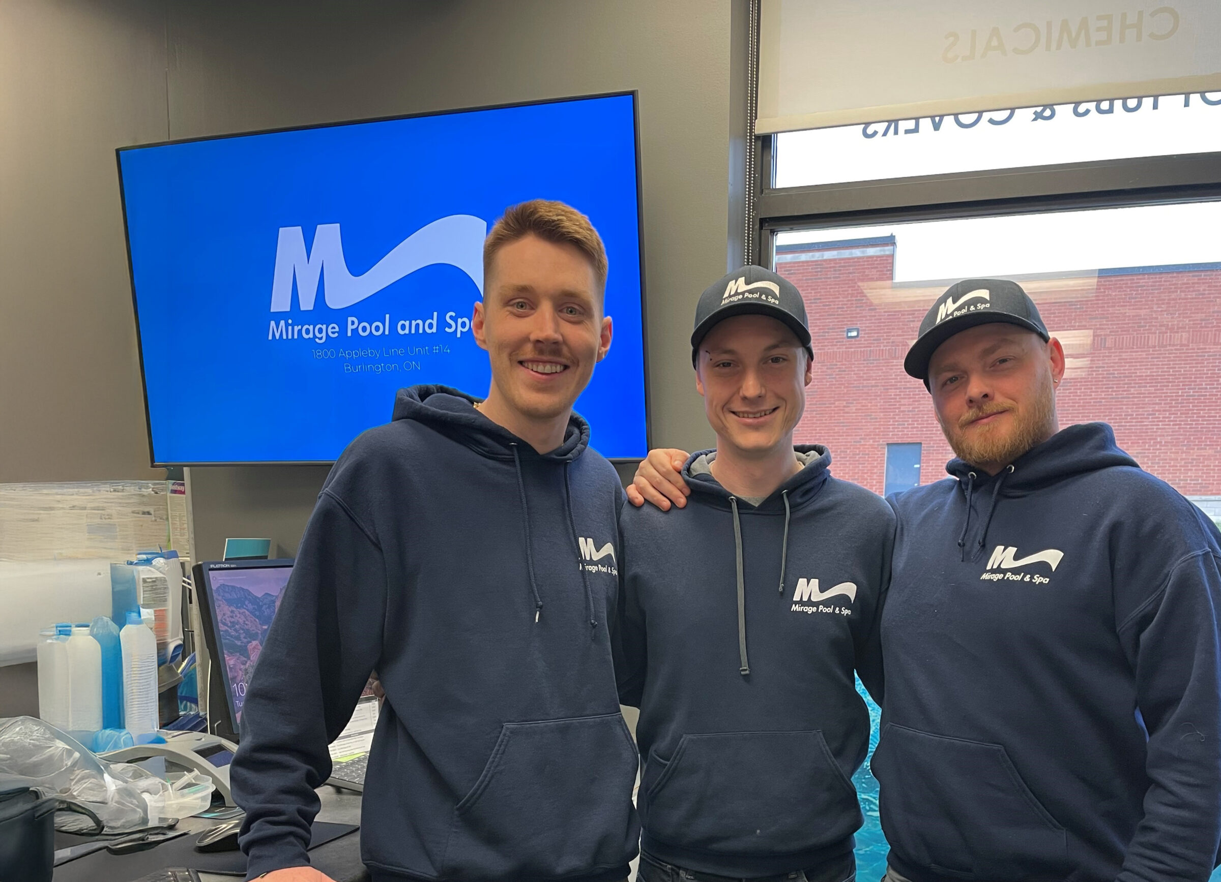 Three people in matching hoodies and caps, smiling indoors near a screen displaying "Mirage Pool and Spa" with a visible outside brick building.