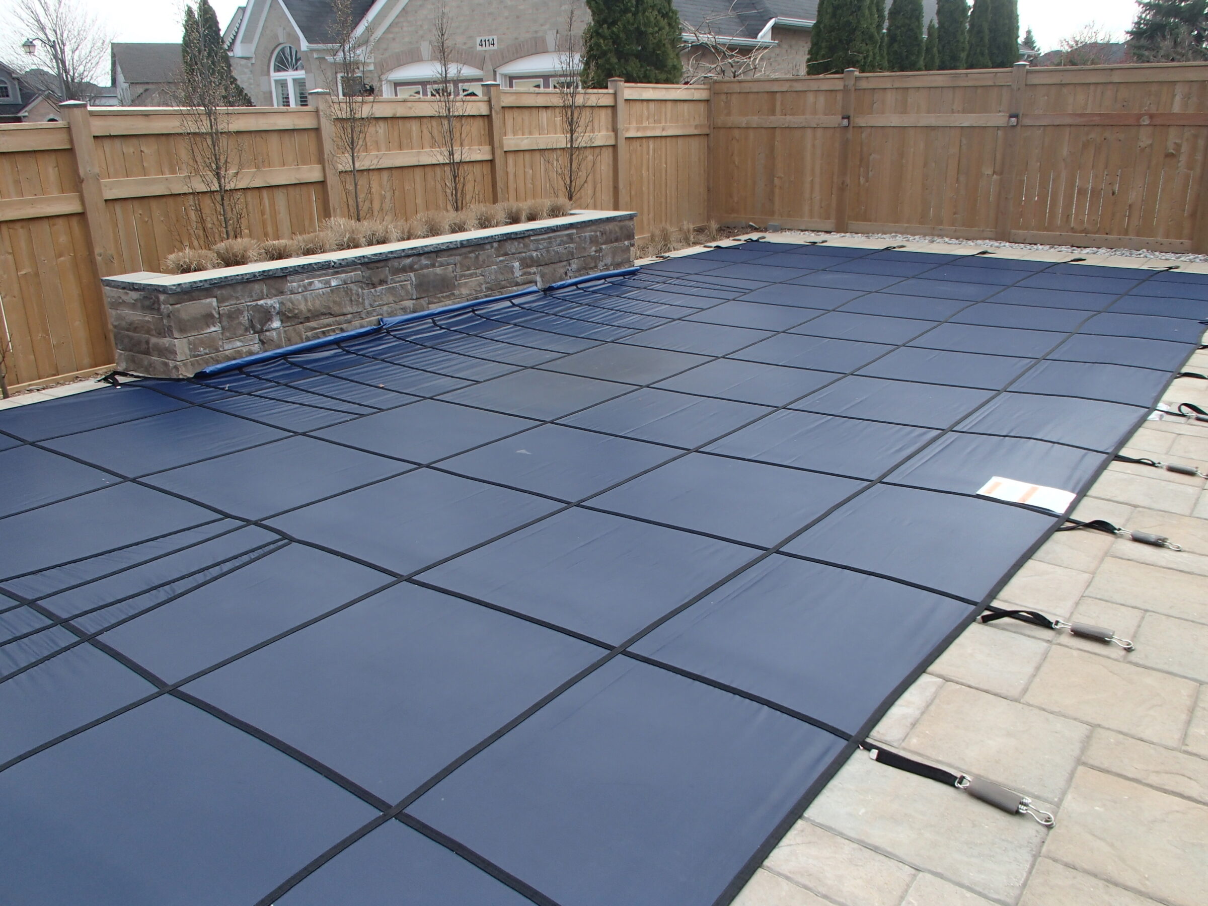 Backyard with a securely covered swimming pool. Wooden fence surrounds the area, and a stone planter is adjacent. Houses and trees are visible.