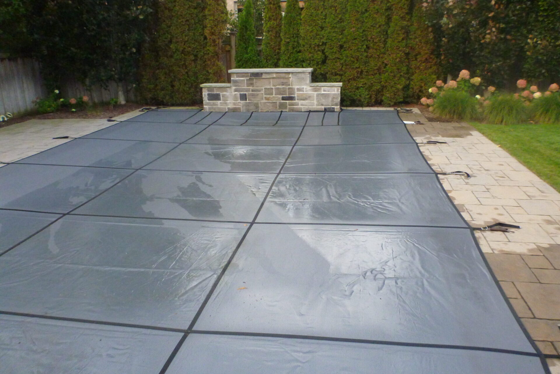 A covered pool in a backyard, surrounded by stone patio and greenery, with a small brick wall and plants in the background.