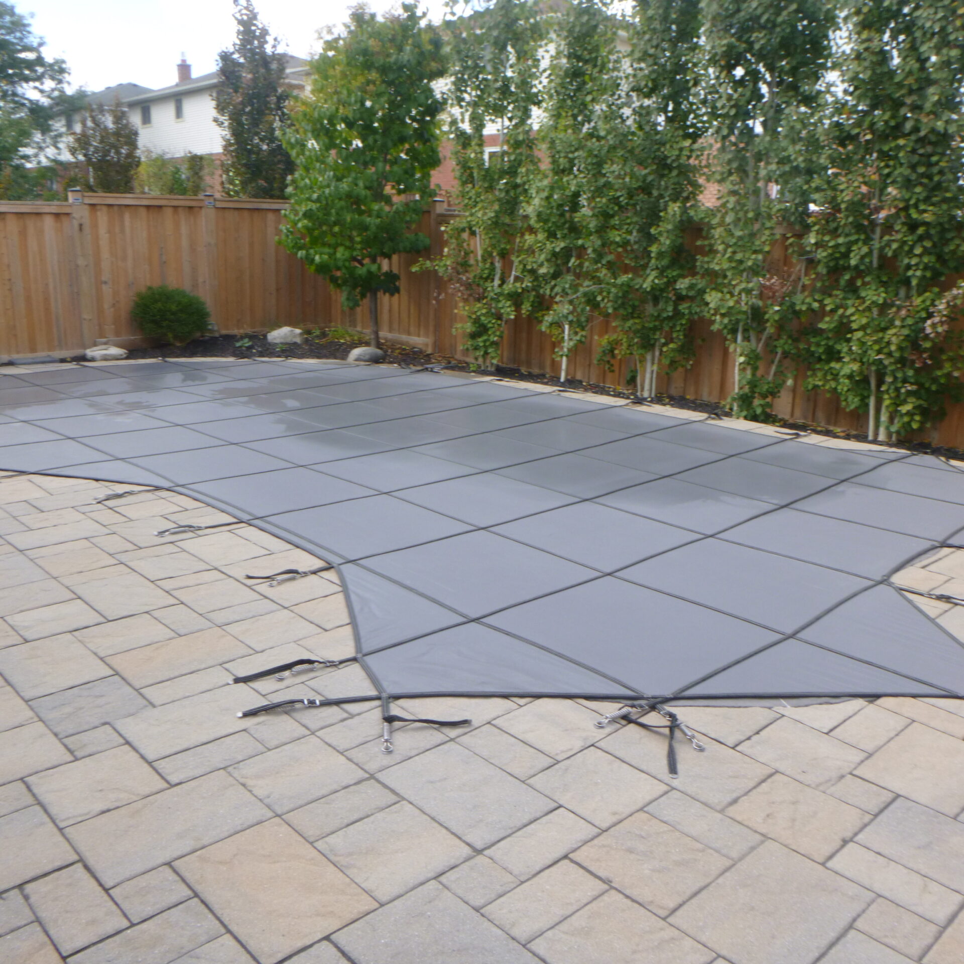 A fenced backyard features a rectangular covered pool surrounded by stone tiles, trees, and shrubs, suggesting a serene, private outdoor space.