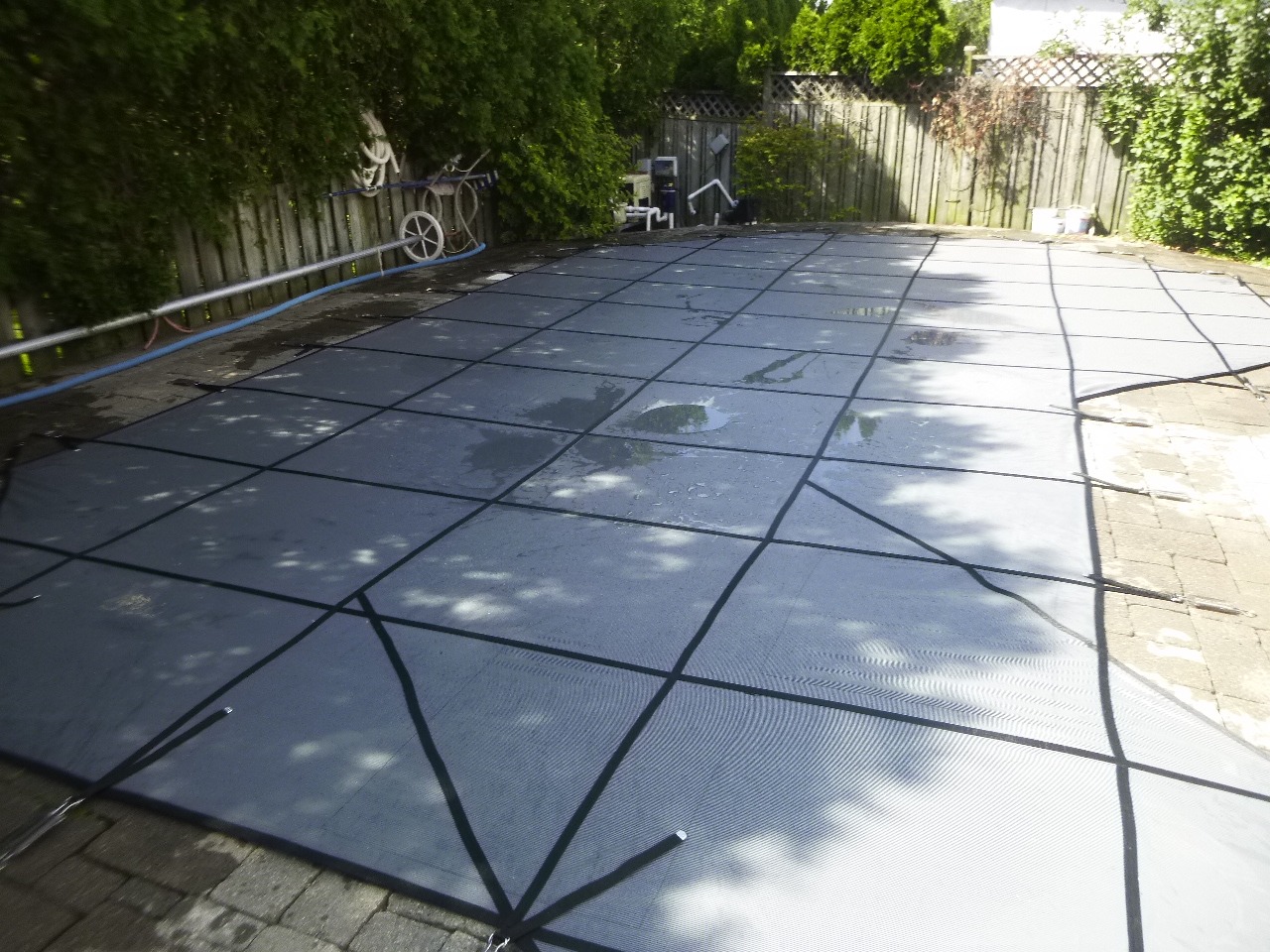 A fenced backyard features a swimming pool covered with a mesh safety cover. Various pool equipment is visible near the wooden fence.