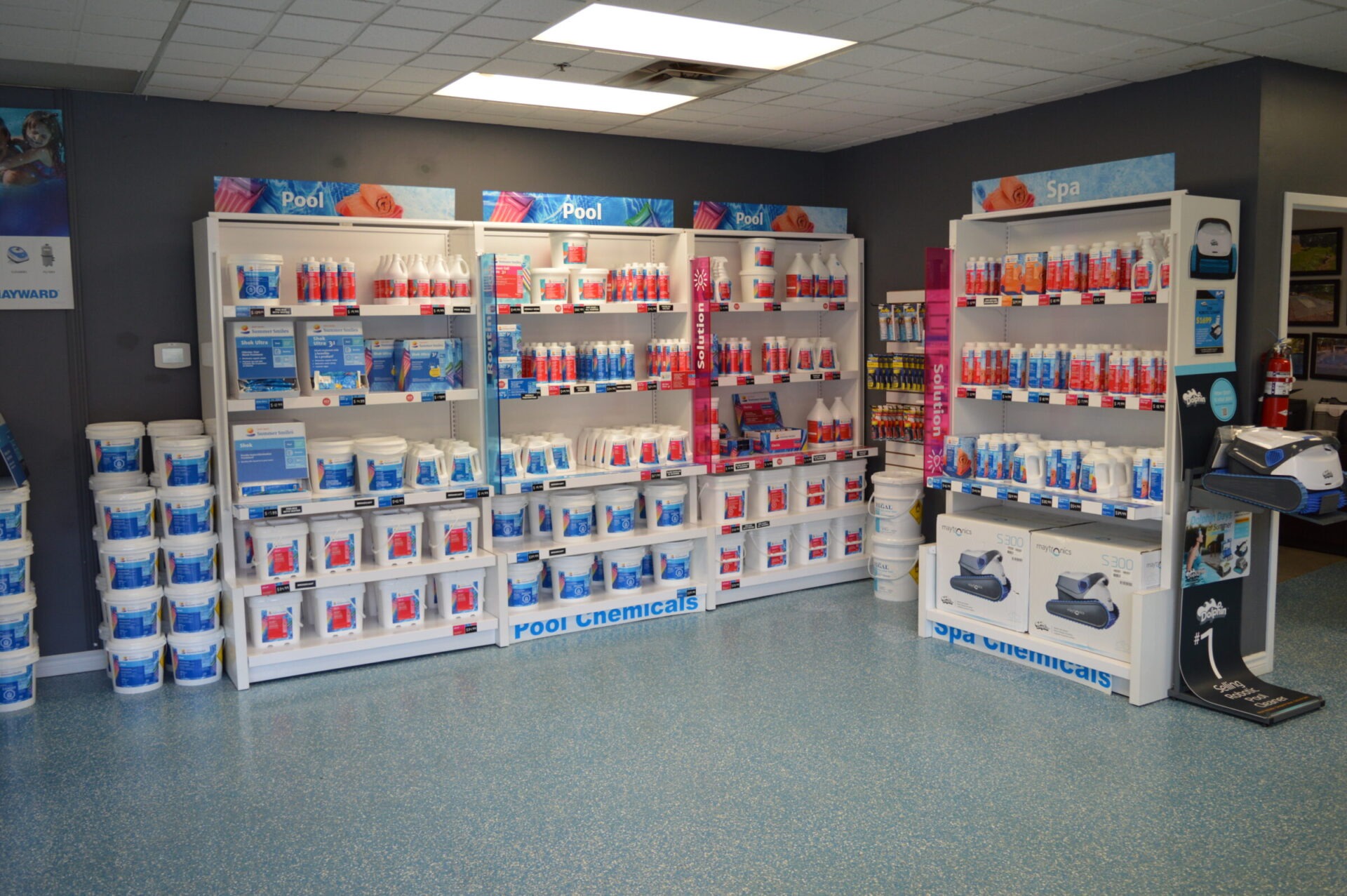 The image shows a store interior with shelves displaying pool and spa chemicals, cleaning products, and supplies against a gray wall.