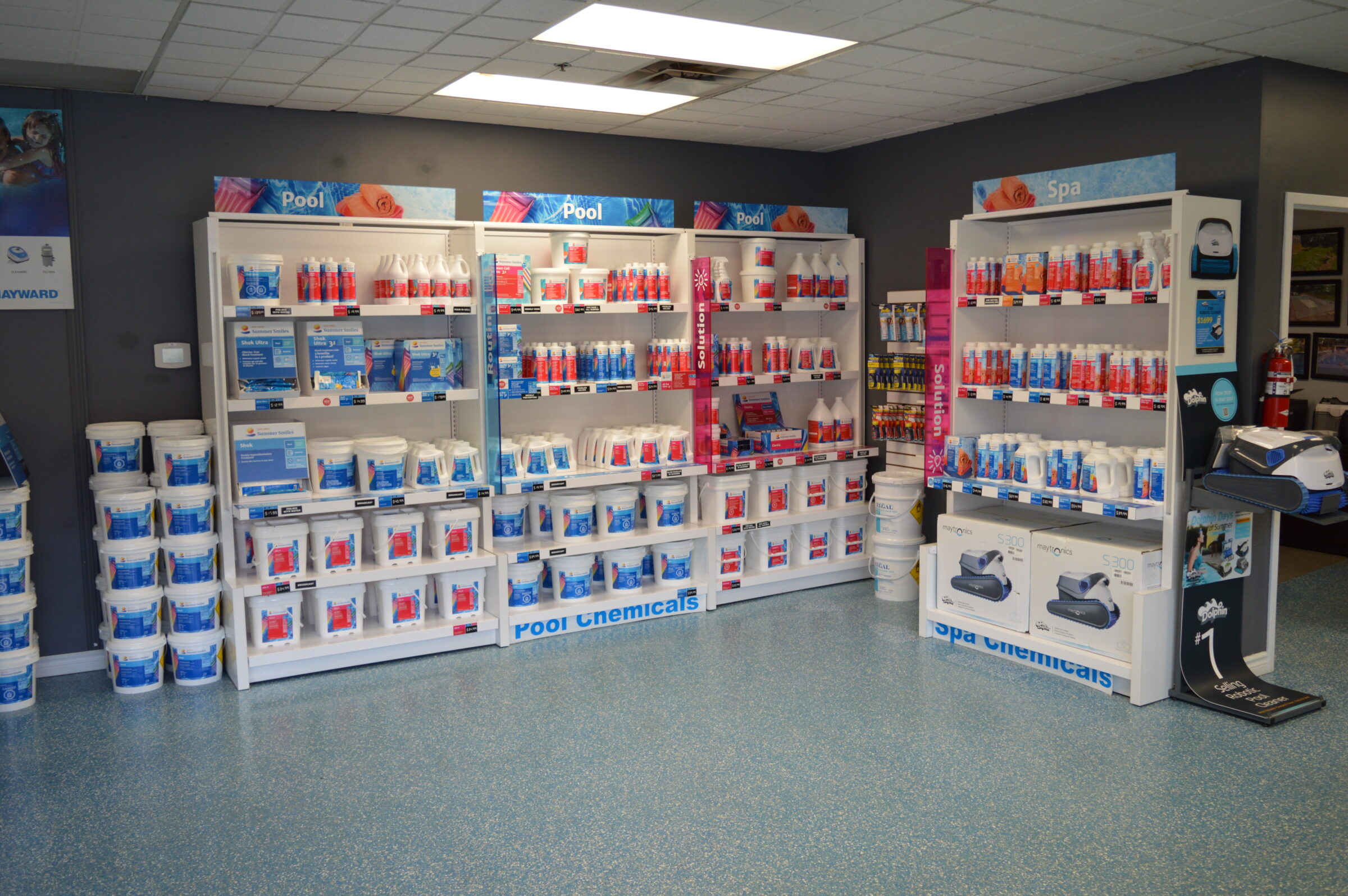 The image shows a store interior with shelves displaying pool and spa chemicals, cleaning products, and supplies against a gray wall.
