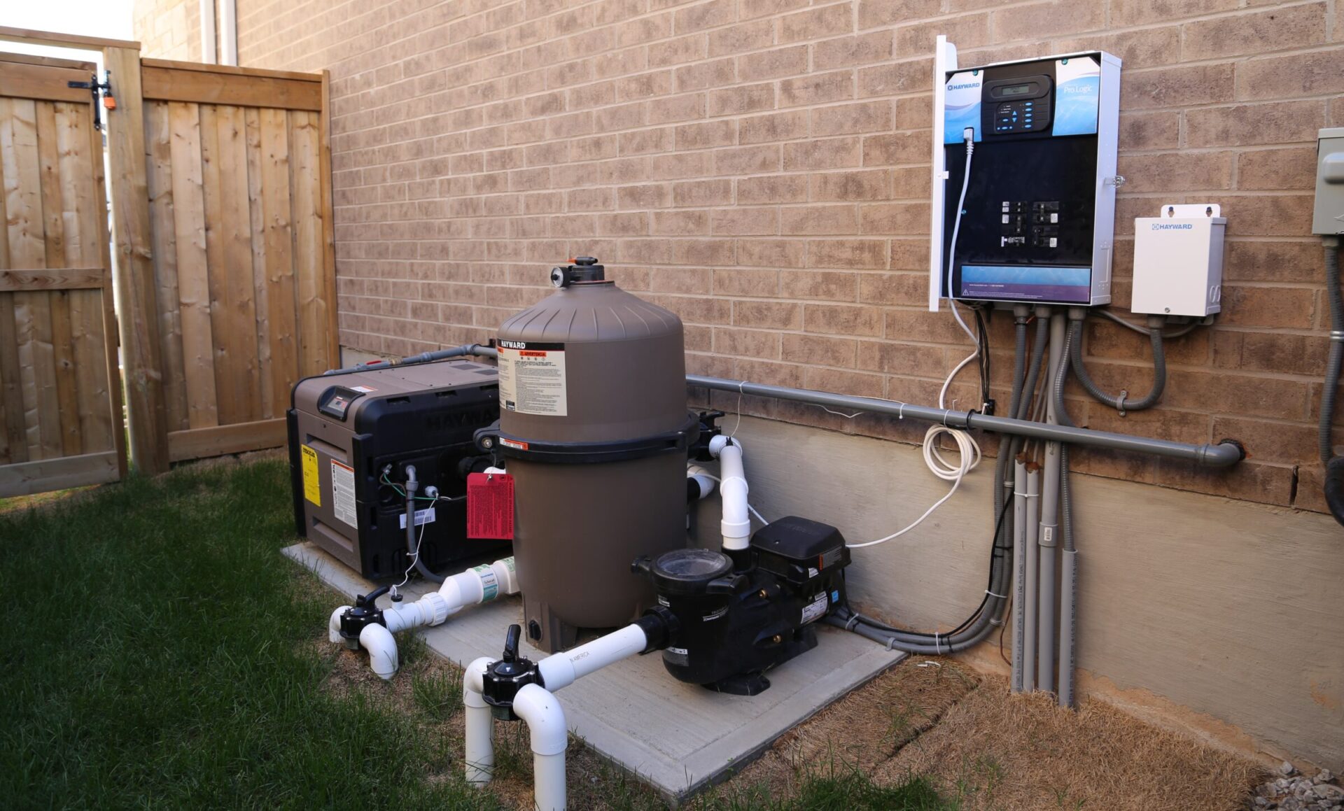 Outdoor pool equipment setup with a filter, pump, heater, and electrical control panel against a brick wall. Grass and wooden fence nearby.